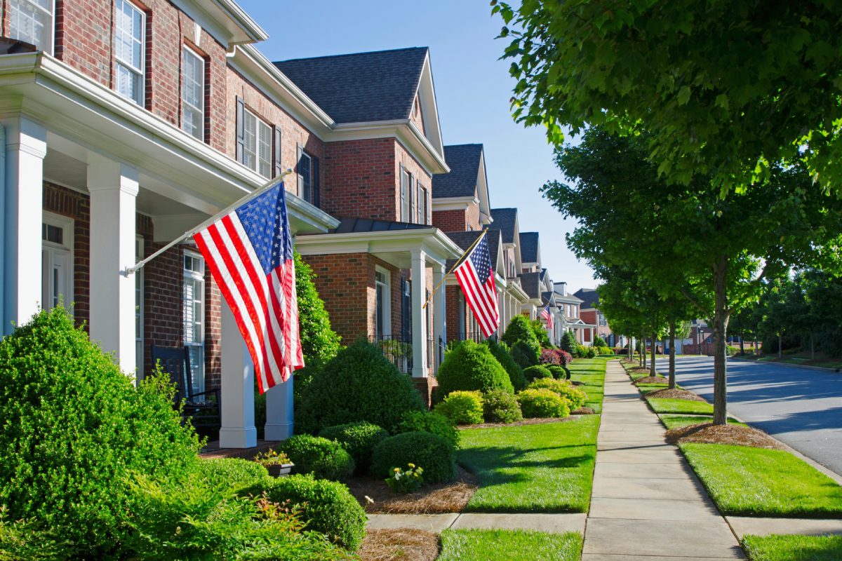 Row of suburban homes with American flags on porches on a sunny day.