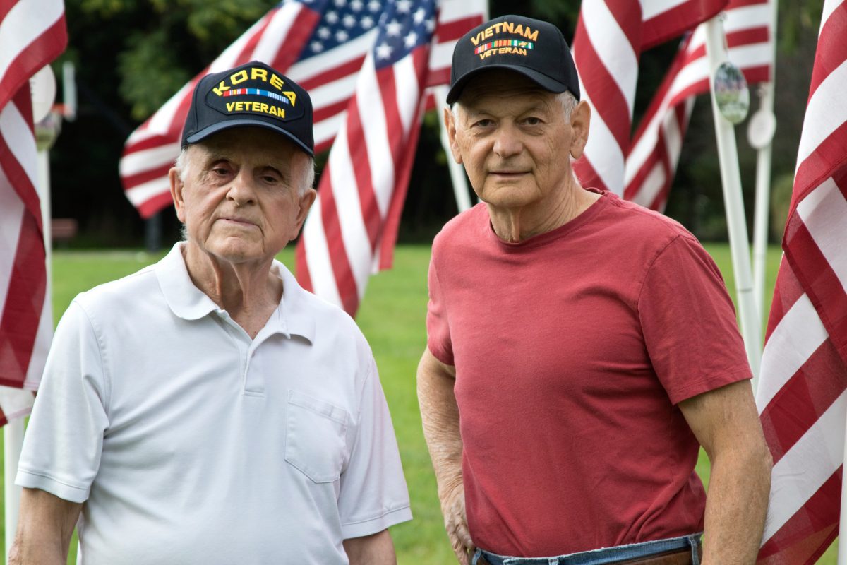 Two older men wearing military caps standing in front of American flags.
