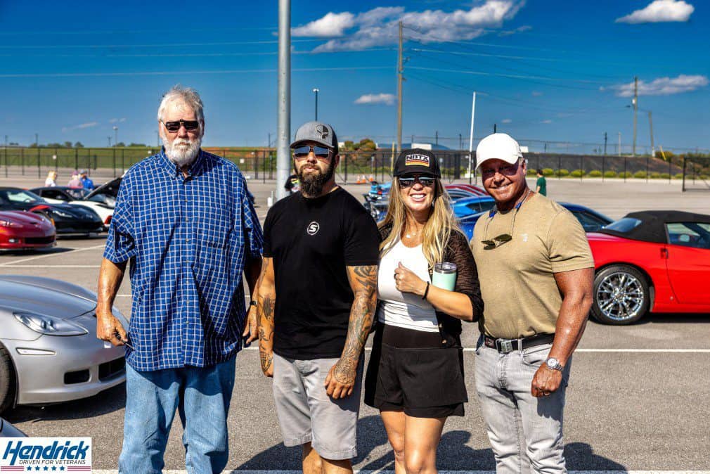 Four people pose among cars at a Hendrick Driven for Veterans event.