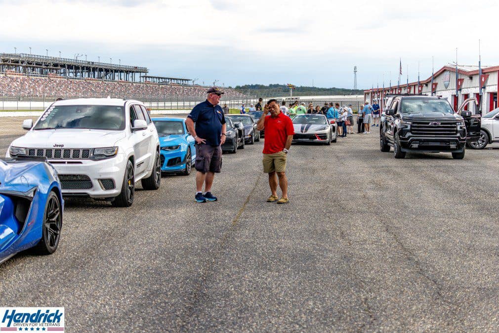 Cars and attendees gather at a racetrack for a Hendrick Driven for Veterans event.