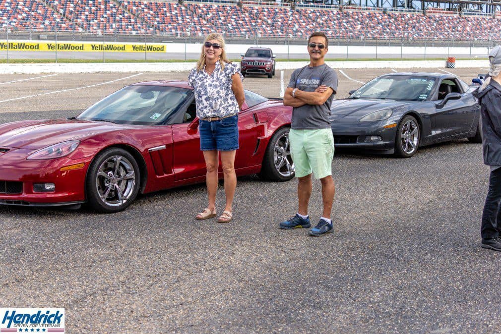 Veterans charity supporters pose beside sports cars at a racetrack fundraising event.